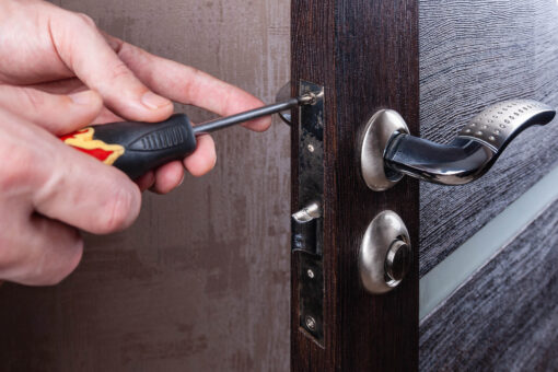 The carpenter installs and adjusts the steel door lock in a wooden casement. Using a screwdriver, a locksmith replaces the old door lock mechanism in the living room.